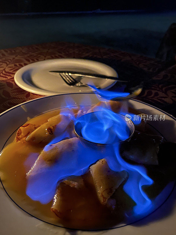 Close-up image of plate of French crêpes (thin pancakes) covered in orange and caramel sauce, Crepe suzette flambe with orange liqueur, blue flame in metal bowl of alcohol, focus on foreground, copy space